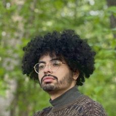 Color headshot of a person with curly hair in a wooded area.