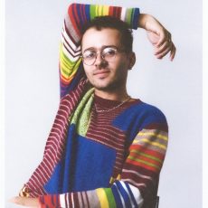 A color headshot of a person wearing a multi-colored sweater looking at the camera.